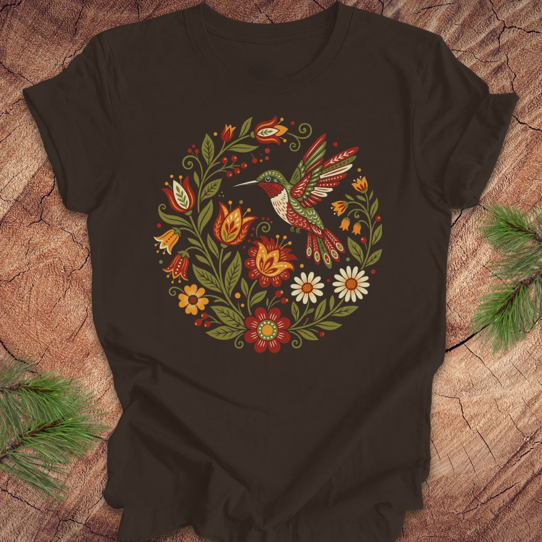 A brown short-sleeve t-shirt with a colorful folk-art style graphic of a hummingbird and flowers, laid flat on a rustic wood background with pine branches in the corners.
