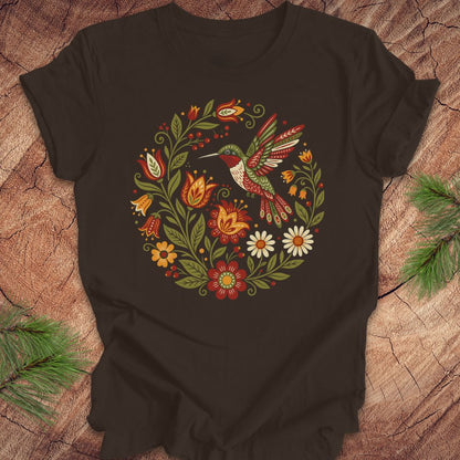 A brown short-sleeve t-shirt with a colorful folk-art style graphic of a hummingbird and flowers, laid flat on a rustic wood background with pine branches in the corners.