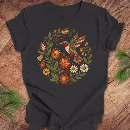 A dark charcoal gray t-shirt featuring a colorful folk-art style graphic of a hummingbird surrounded by flowers and leaves, laid flat on a rustic wood background with pine needles in the corners.