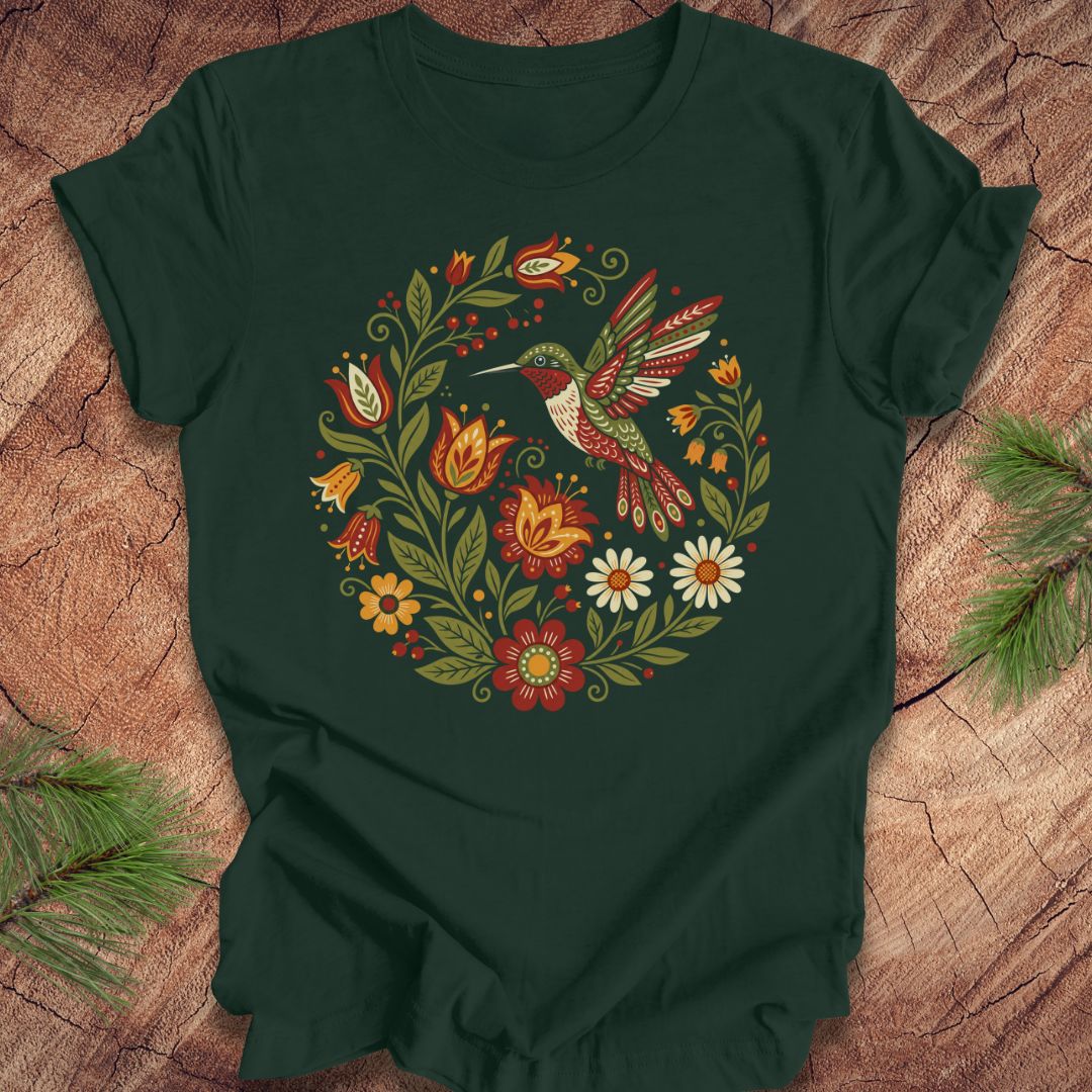 A forest green short-sleeve t-shirt with a colorful folk-art style graphic of a hummingbird and flowers, laid flat on a rustic wood background with pine branches in the corners.