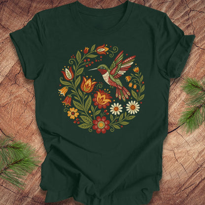 A forest green short-sleeve t-shirt with a colorful folk-art style graphic of a hummingbird and flowers, laid flat on a rustic wood background with pine branches in the corners.