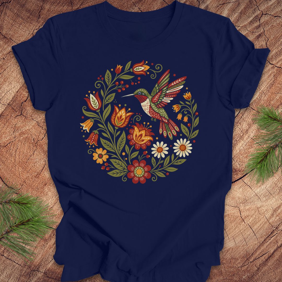 A navy blue short-sleeve t-shirt with a colorful folk-art style graphic of a hummingbird and flowers, laid flat on a rustic wood background with pine branches in the corners.