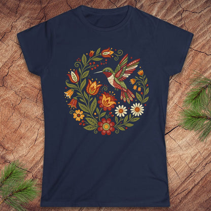 A navy blue short-sleeve crewneck t-shirt featuring a colorful circular graphic of a hummingbird surrounded by flowers, laid flat on a textured wood background with pine needles in the corners.