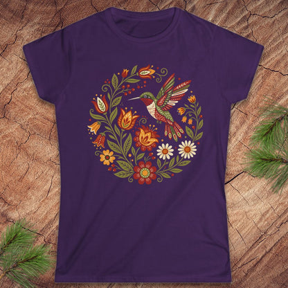 A purple short-sleeve crewneck t-shirt featuring a colorful graphic of a hummingbird surrounded by a circular floral wreath, laid flat on a textured wood background with pine needles in the corners.