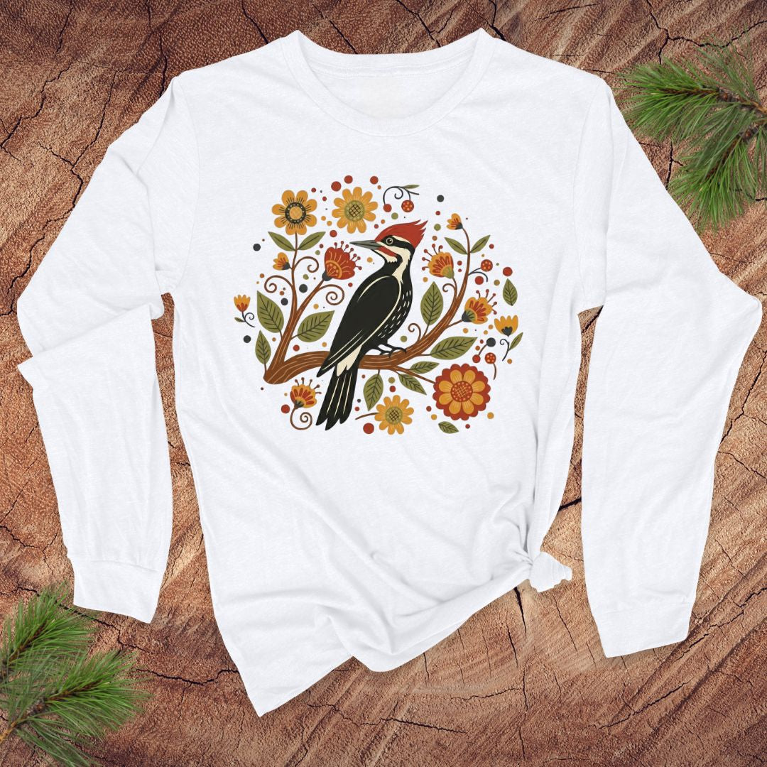 White long sleeve t-Shirt with a pileated woodpecker on a branch surrounded by folk art flowers on a wood background.