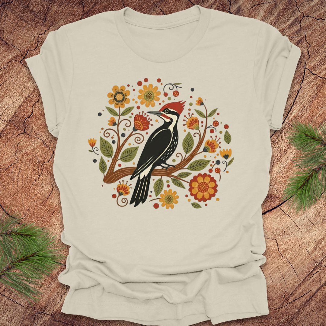 Beige T-Shirt with a pileated woodpecker on a branch surrounded by folk art flowers on a wood background.