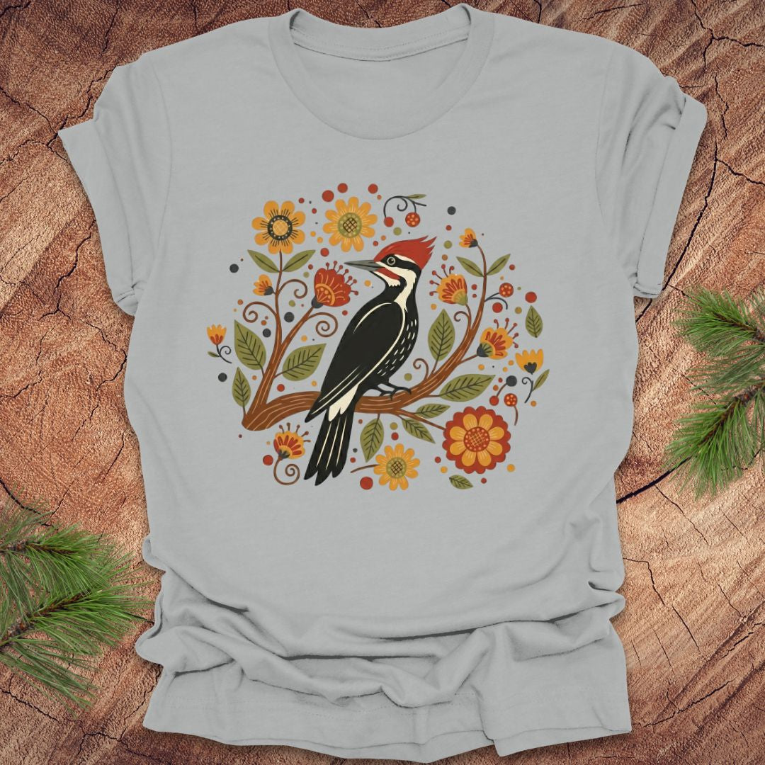 Silver T-Shirt with a pileated woodpecker on a branch surrounded by folk art flowers on a wood background.