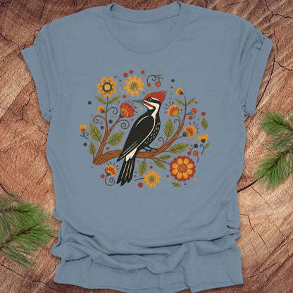 Steel blue T-Shirt with a pileated woodpecker on a branch surrounded by folk art flowers on a wood background.