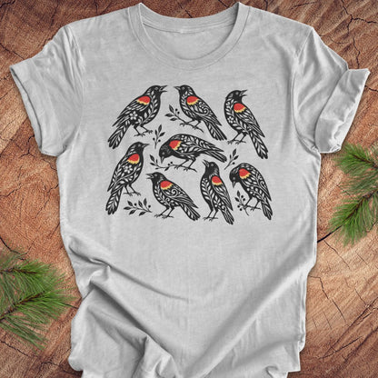 A light gray crewneck t-shirt featuring a graphic print of nine red-winged blackbirds with red chest patches, laid flat on a rustic wood background with pine branches.