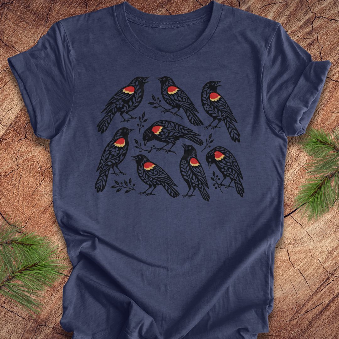 A navy blue short-sleeve t-shirt with a graphic print of nine black birds featuring red and yellow chest patches, laid flat on a rustic wood background with pine branches in the corners.