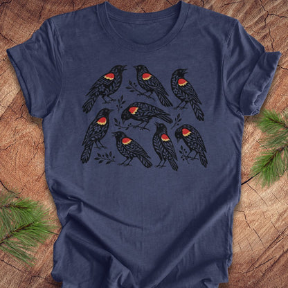 A navy blue short-sleeve t-shirt with a graphic print of nine black birds featuring red and yellow chest patches, laid flat on a rustic wood background with pine branches in the corners.
