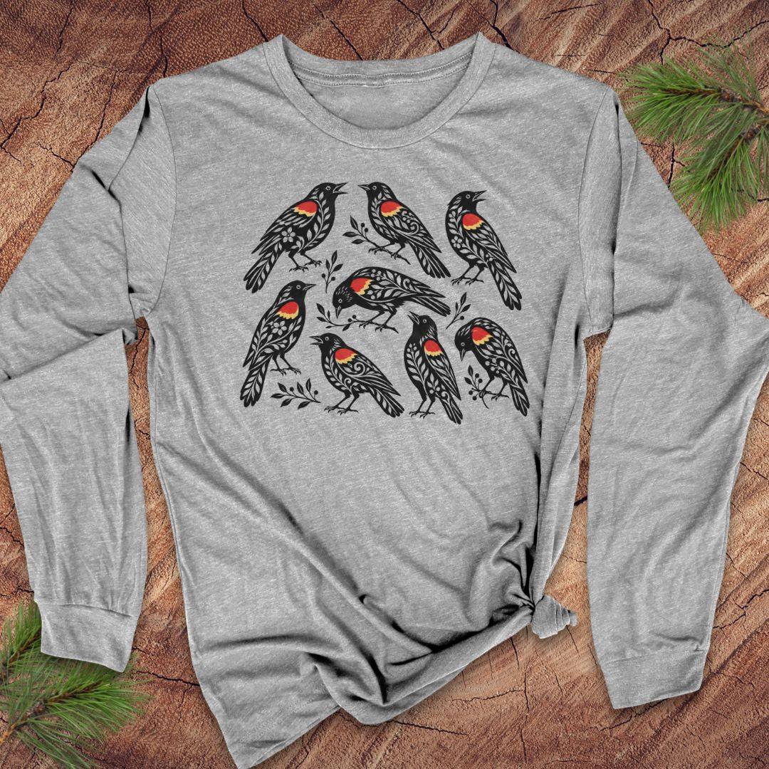 A heather gray long-sleeve t-shirt with a graphic print of seven red-winged blackbirds, laid flat on a rustic wood background with pine needles.