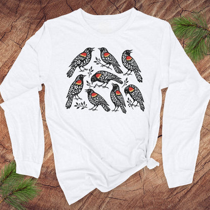 A white long-sleeve t-shirt with a graphic of seven red-winged blackbirds, laid flat on a rustic wood background with pine needles in the corners.