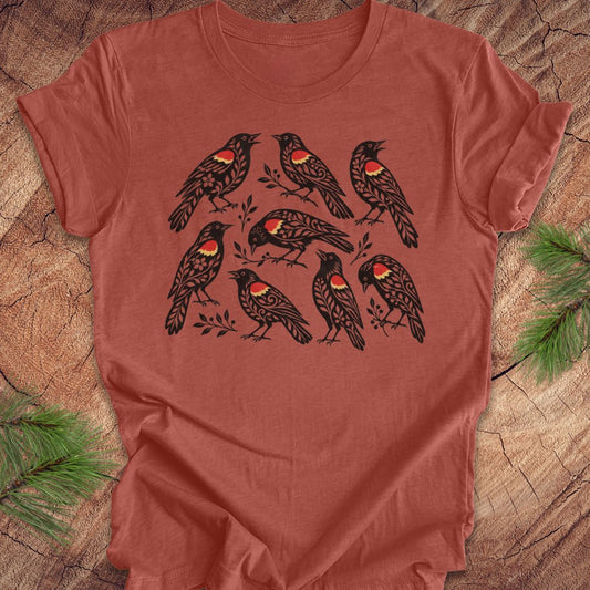 A terracotta-colored short-sleeve t-shirt featuring a graphic print of nine black birds with red and yellow chest details, laid flat on a rustic wood background with pine branches.