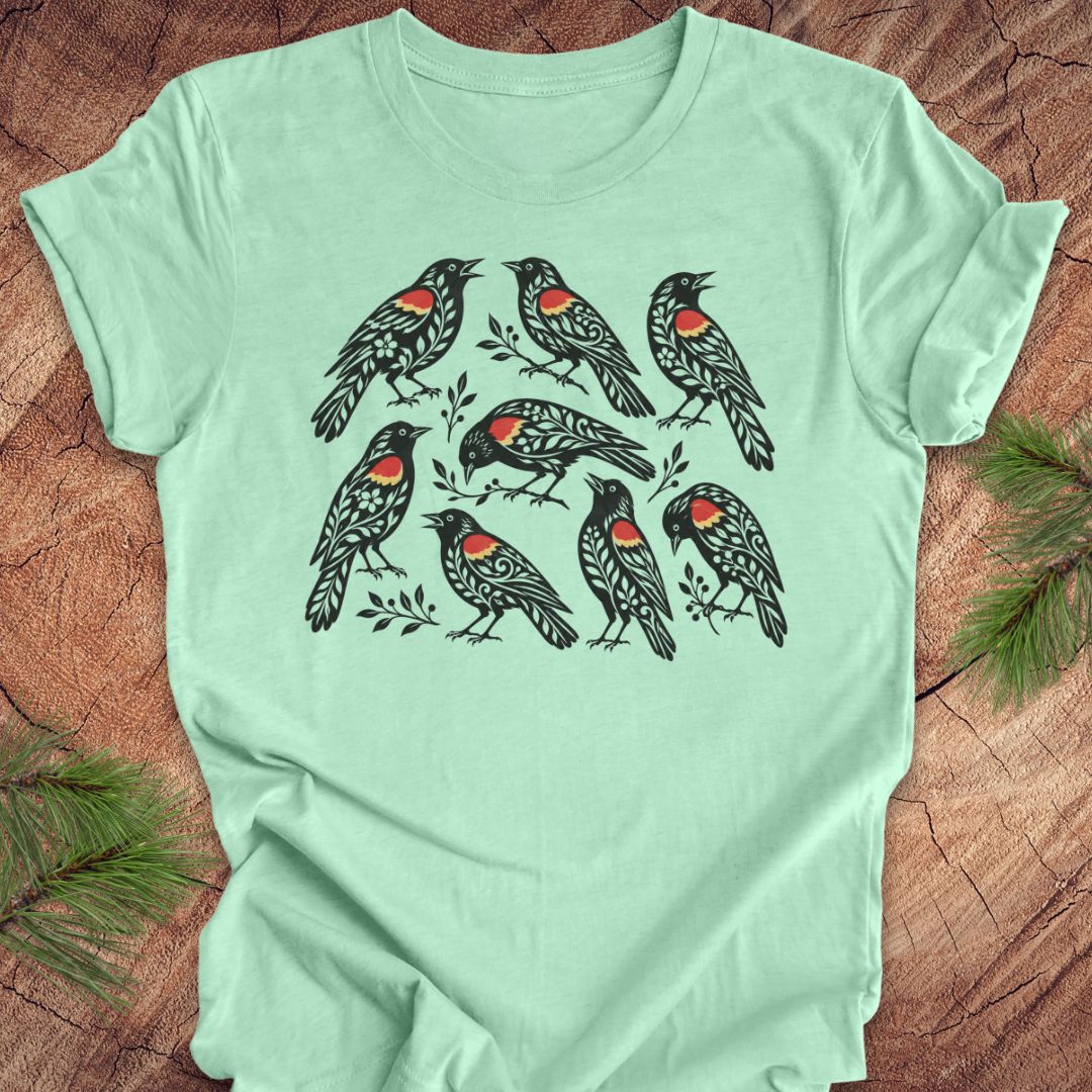 A mint green short-sleeve t-shirt with a graphic print of seven red-winged blackbirds, laid flat on a rustic wood background with pine branches in the corners.