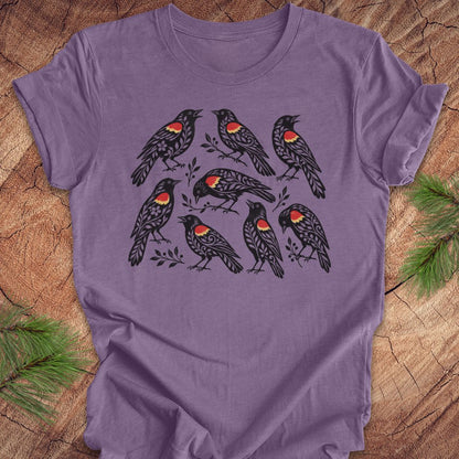 A purple short-sleeve crewneck t-shirt featuring a graphic print of eight black birds with red and yellow chest patches, laid flat on a rustic wood background with pine branches in the corners.