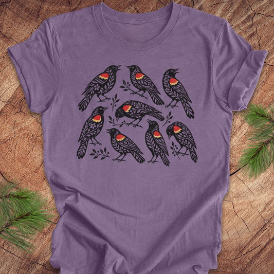 A purple short-sleeve crewneck t-shirt featuring a graphic print of eight black birds with red and yellow chest patches, laid flat on a rustic wood background with pine branches in the corners.