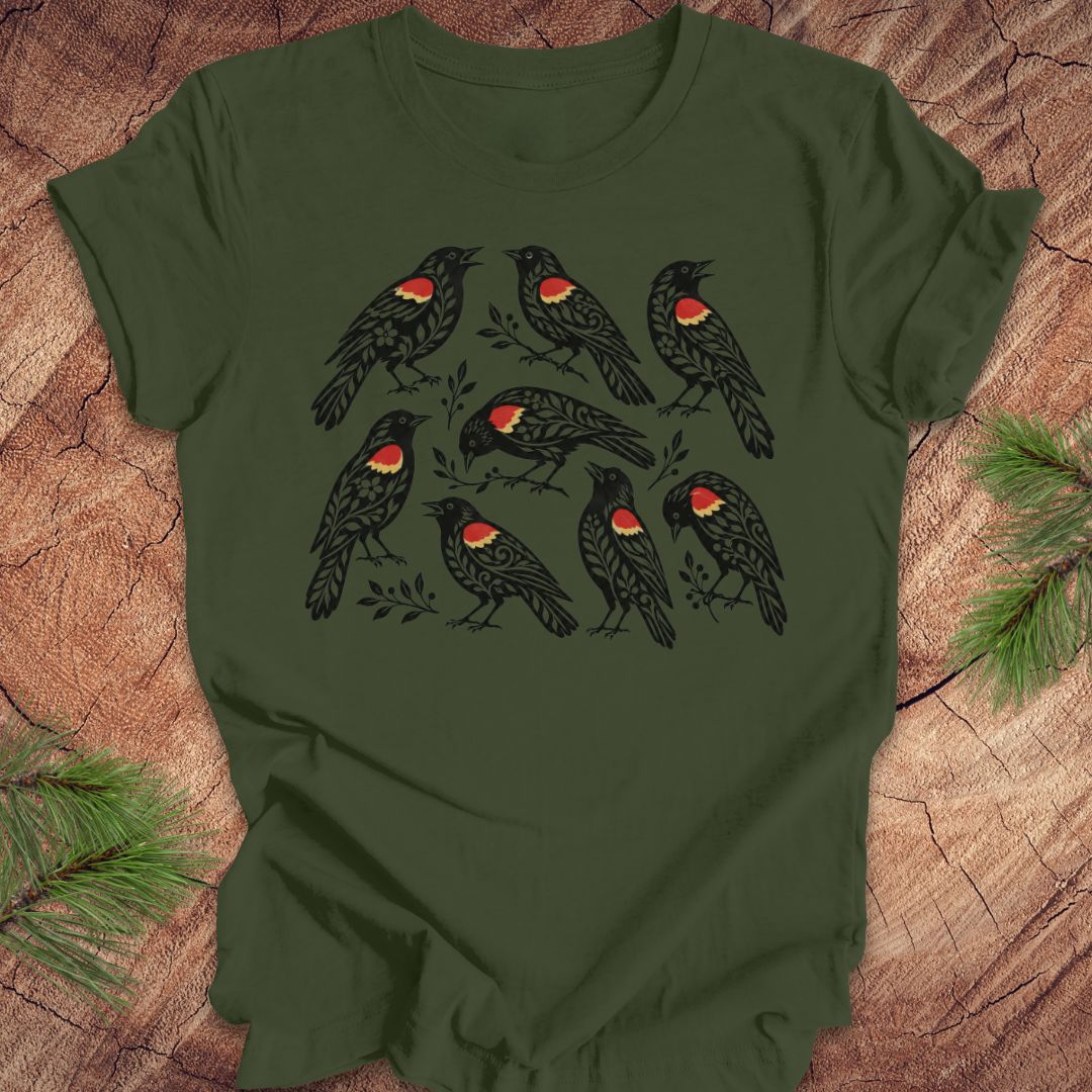 An olive green short-sleeve t-shirt featuring a graphic print of nine black birds with red and yellow chest patches, laid flat on a rustic wood background with pine branches in the corners.