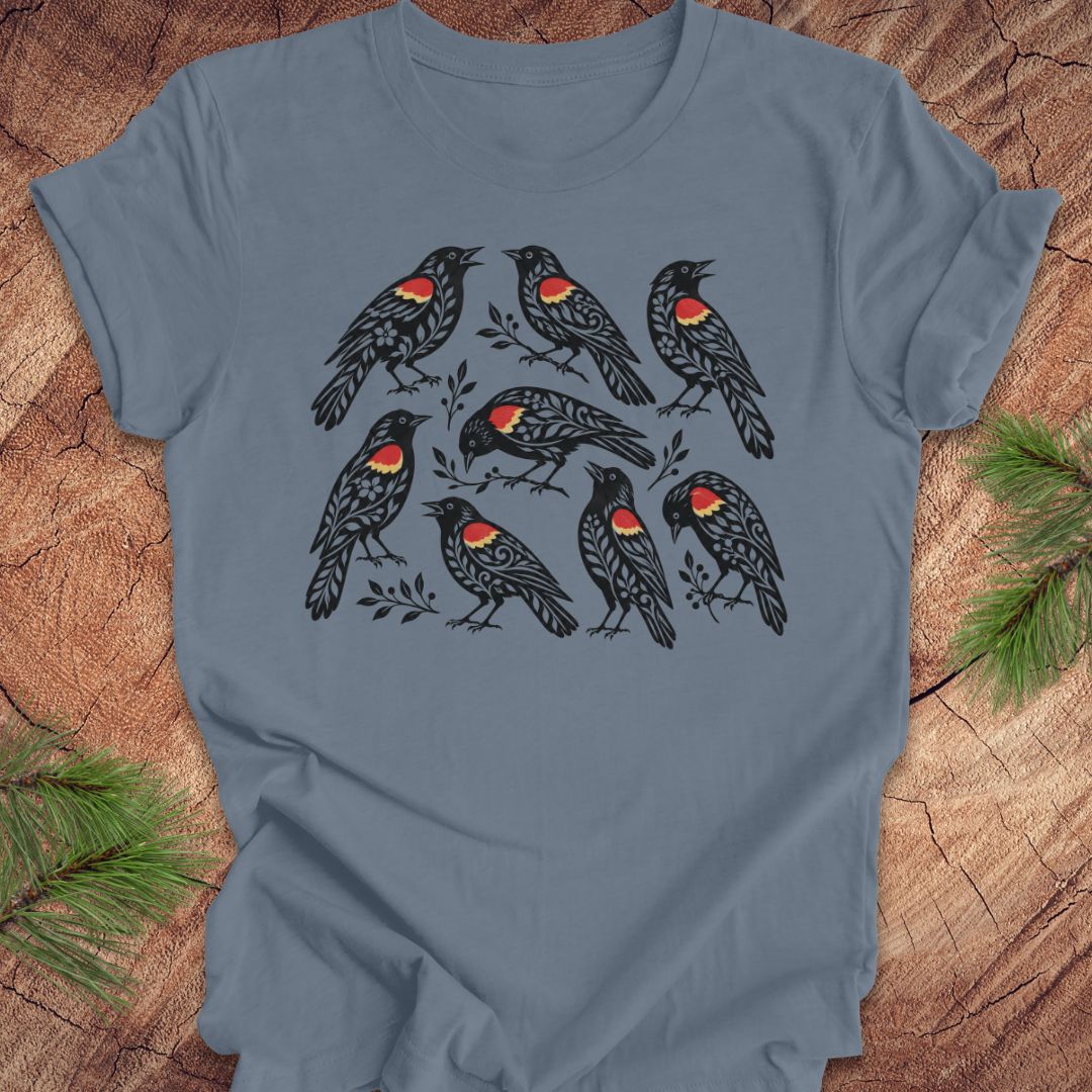 A slate blue short-sleeve t-shirt with a graphic print of nine black birds featuring red and yellow chest patches, displayed flat on a rustic wood background with pine branches.