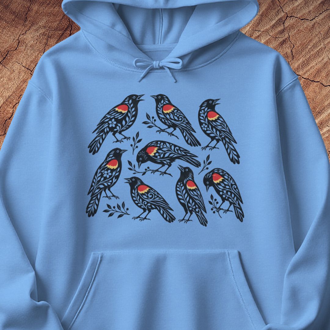 A light blue pullover hoodie laid flat on a rustic wood background, featuring a graphic print of eight black birds with red and yellow chest details.