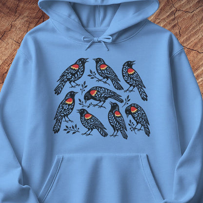 A light blue pullover hoodie laid flat on a rustic wood background, featuring a graphic print of eight black birds with red and yellow chest details.