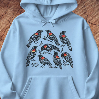 A light blue pullover hoodie featuring a graphic print of eight black birds with red and orange chest patches, laid flat on a textured brown wood background.