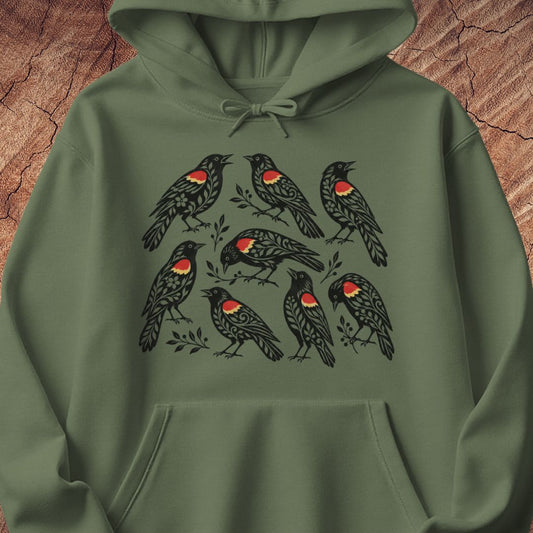 An green hoodie featuring a graphic print of eight red-winged blackbirds with red chest patches, displayed against a rustic wood background.