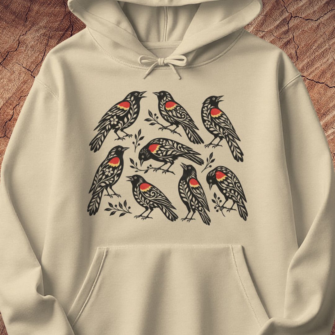 An off-white hoodie featuring a graphic print of eight red-winged blackbirds with red chest patches, displayed against a rustic wood background.