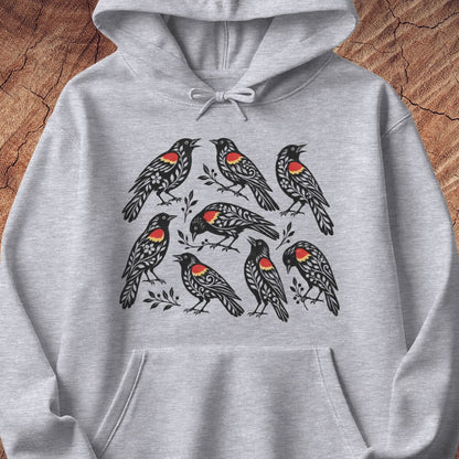 A light heather gray pullover hoodie featuring a graphic print of seven red-winged blackbirds with bright red and orange chest patches, displayed flat against a rustic wood background.