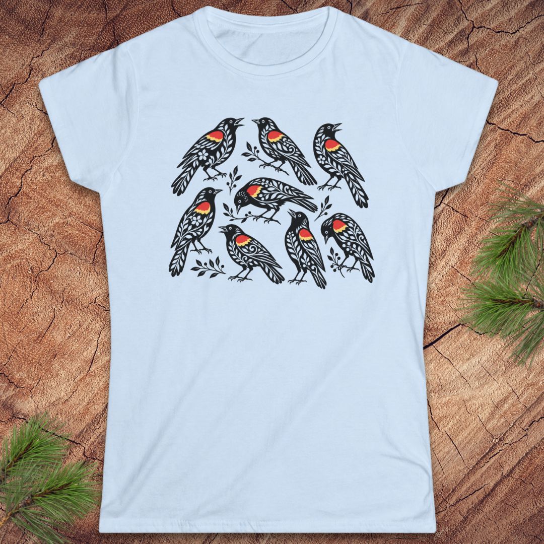A light blue short-sleeve crewneck t-shirt featuring a graphic print of seven red-winged blackbirds on branches, laid flat on a rustic wood background with pine needles in the corners.