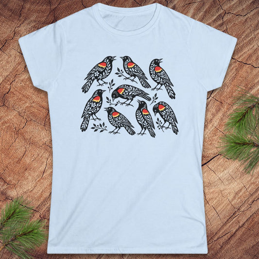 A light blue short-sleeve crewneck t-shirt featuring a graphic print of seven red-winged blackbirds on branches, laid flat on a rustic wood background with pine needles in the corners.