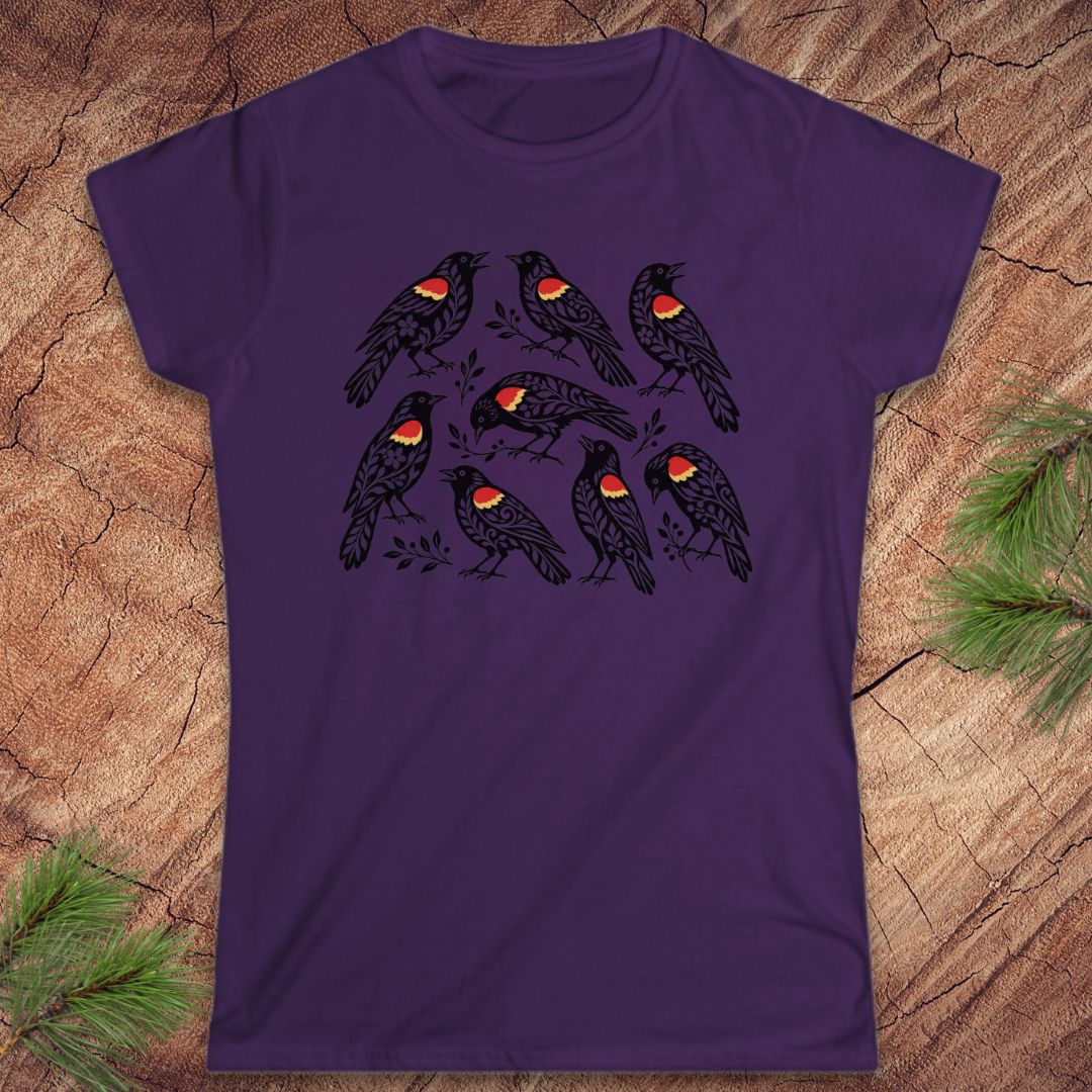 A purple short-sleeve crewneck t-shirt featuring a graphic print of eight black birds with red and yellow wattles, laid flat on a rustic wood background with pine needles in the corners.