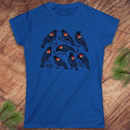A royal blue short-sleeve crewneck t-shirt featuring a graphic print of seven red-winged blackbirds perched on branches. The t-shirt is displayed flat on a textured wood background with pine needles in the corners.