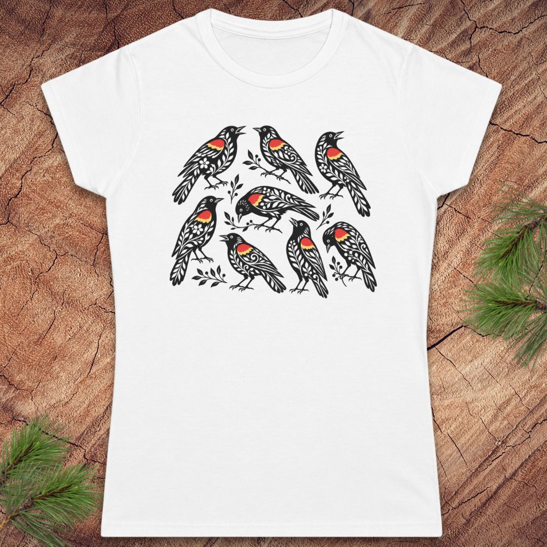 A white short-sleeve crewneck t-shirt featuring a graphic print of nine black birds with red and orange chest patches, laid flat on a rustic wood background with pine needles in the corners.