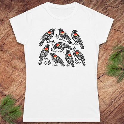 A white short-sleeve crewneck t-shirt featuring a graphic print of nine black birds with red and orange chest patches, laid flat on a rustic wood background with pine needles in the corners.