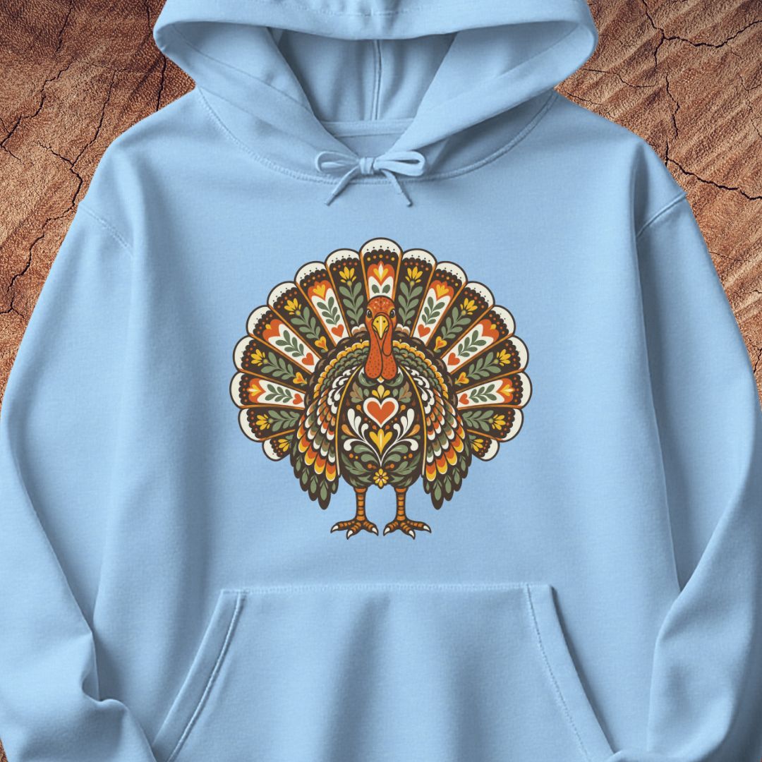 A light blue pullover hoodie featuring a large, colorful, ornate turkey graphic printed on the chest, laid flat on a textured brown wood background.