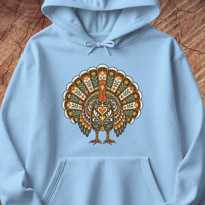A light blue pullover hoodie featuring a large, colorful, ornate turkey graphic printed on the chest, laid flat on a textured brown wood background.