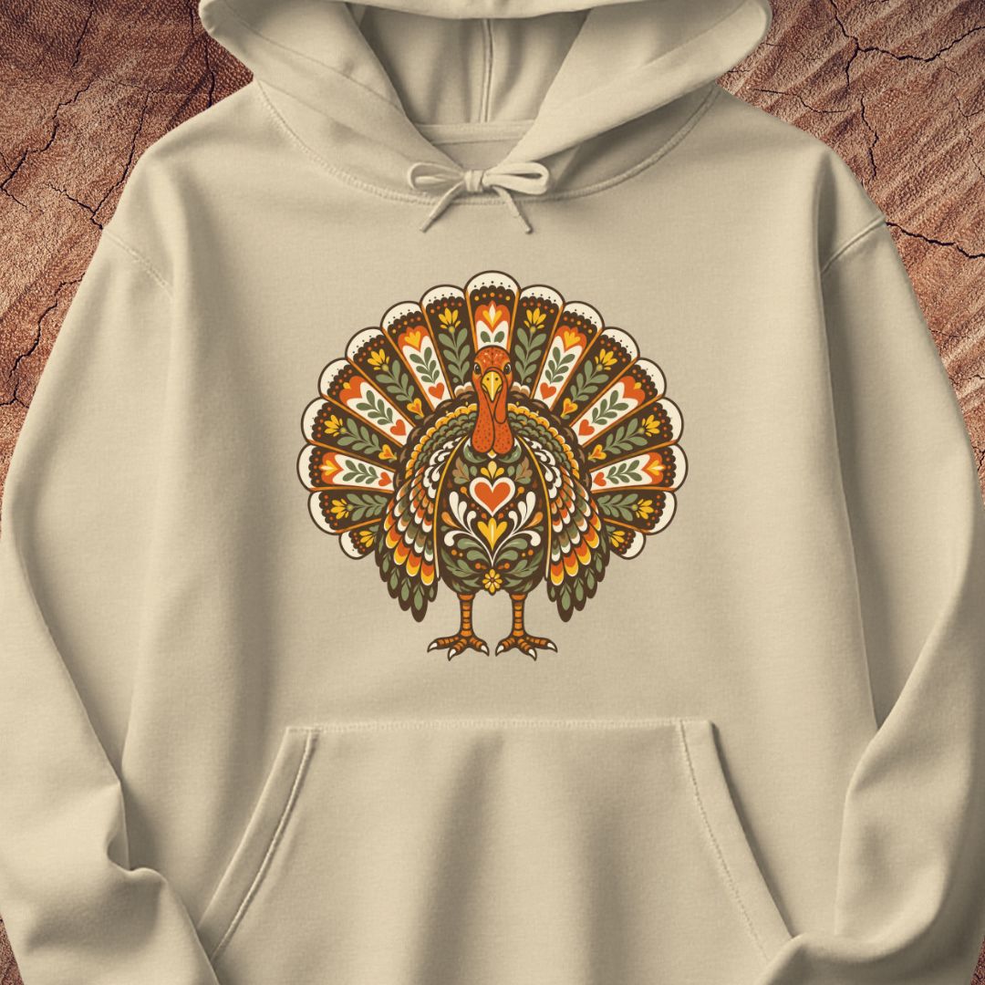 A cream-colored hoodie featuring a detailed, colorful folk-art style turkey graphic on the chest, displayed against a textured brown wood background.