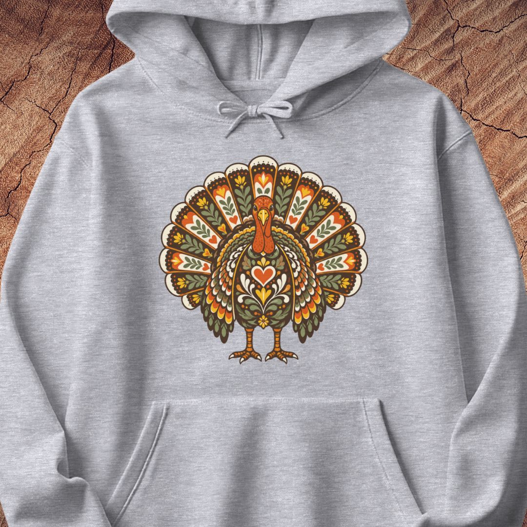 A light gray heather pullover hoodie featuring a large, colorful, ornate graphic of a turkey on the chest, displayed flat against a rustic wood background.