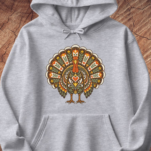 A light gray heather pullover hoodie featuring a large, colorful, ornate graphic of a turkey on the chest, displayed flat against a rustic wood background.