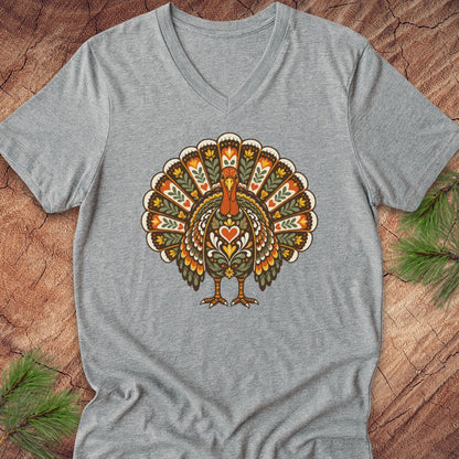 A heather gray V-neck t-shirt featuring a colorful, patterned graphic of a turkey on the chest, laid flat on a rustic wood background with pine needles.