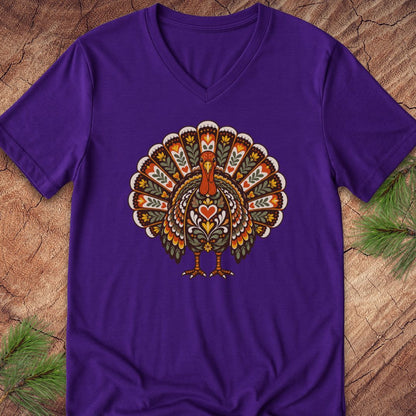 A purple V-neck t-shirt featuring a detailed, colorful Thanksgiving turkey graphic on the chest, laid flat on a rustic wood background with pine needles in the corners.
