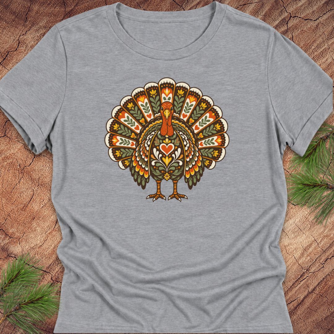 A heather gray short-sleeve t-shirt featuring a colorful, folk-art style turkey graphic on the chest, laid flat on a rustic wood background with pine needles in the corners.