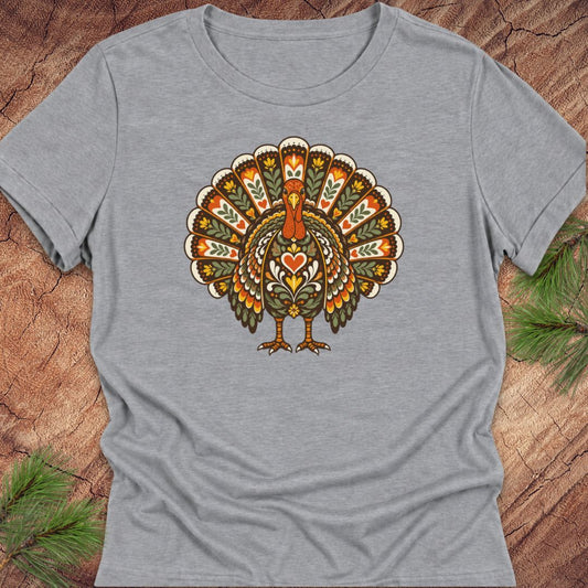 A heather gray short-sleeve t-shirt featuring a colorful, folk-art style turkey graphic on the chest, laid flat on a rustic wood background with pine needles in the corners.