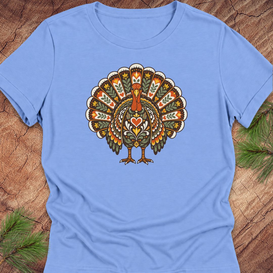 A light blue short-sleeve crewneck t-shirt featuring a detailed, colorful folk-art style turkey graphic on the chest, laid flat on a rustic wood background with pine needles in the corners.
