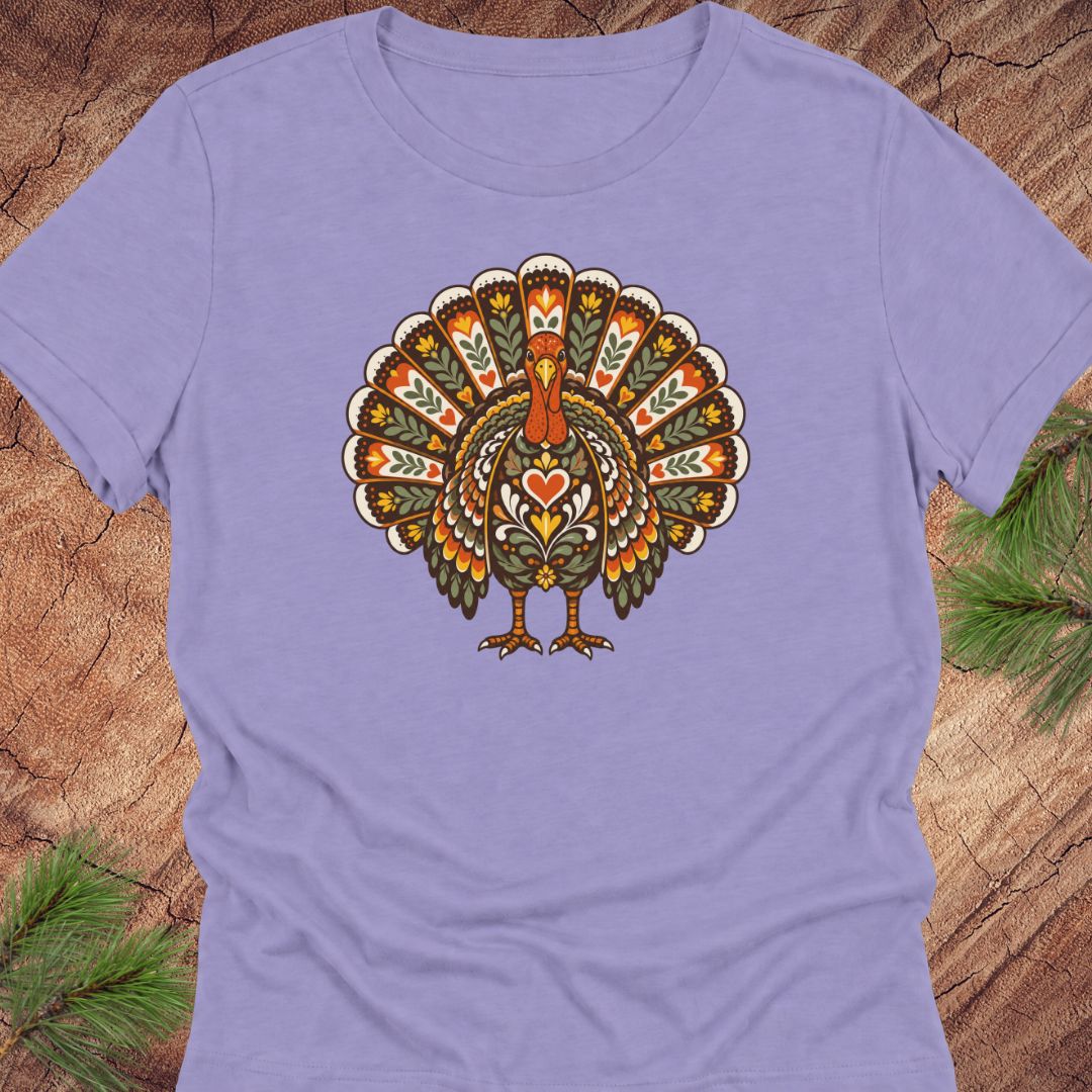 A lavender purple short-sleeve t-shirt featuring a colorful, ornate graphic of a turkey with its tail feathers fanned, displayed on a rustic wood background with pine needles.