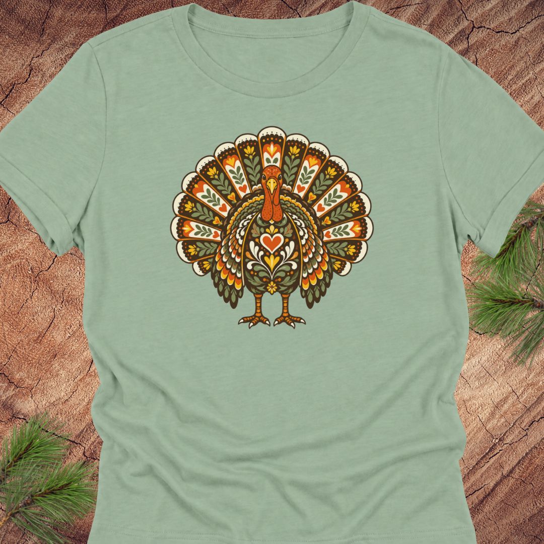 A sage green short-sleeve t-shirt featuring a retro-style graphic of a turkey with a heart on its chest, displayed flat on a rustic wood background with pine needles.
