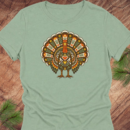 A sage green short-sleeve t-shirt featuring a retro-style graphic of a turkey with a heart on its chest, displayed flat on a rustic wood background with pine needles.