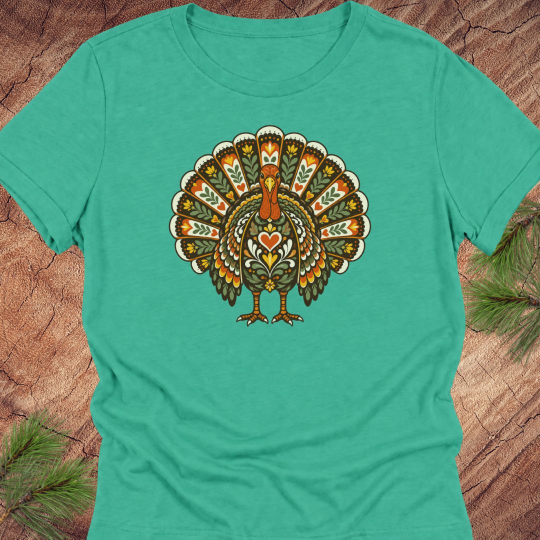 A teal short-sleeve crewneck t-shirt featuring a colorful, ornate graphic of a stylized turkey on the chest, laid flat on a rustic wood background with pine needles in the corners.