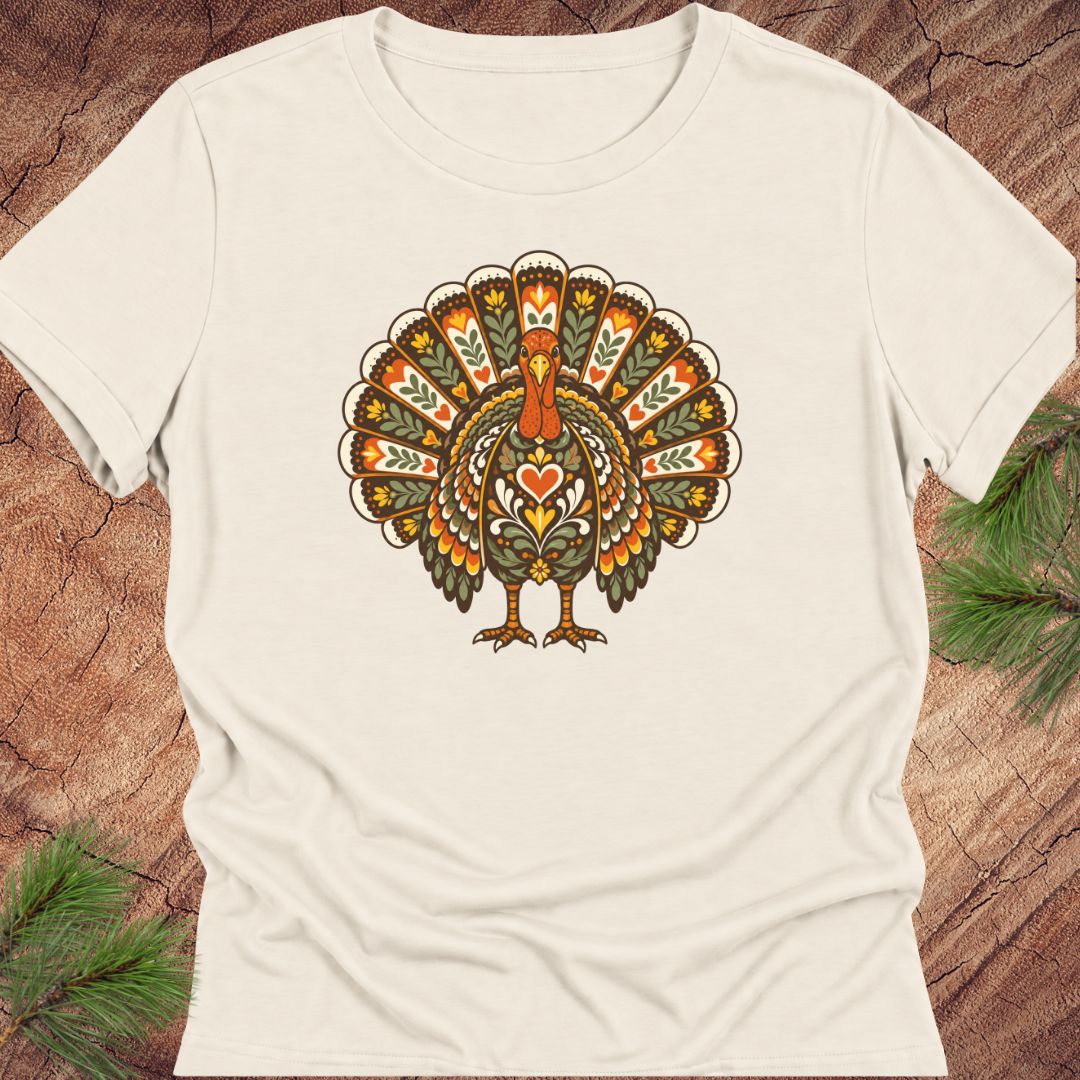 An off-white short-sleeve crewneck t-shirt featuring a colorful, vintage-style graphic of a turkey with a heart on its chest, laid flat on a rustic wood background with pine needles.